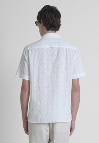 Camicetta bianca a maniche corte con un motivo geometrico testurizzato. Presenta un colletto classico, una fessura sul retro e sottili dettagli cuciti.