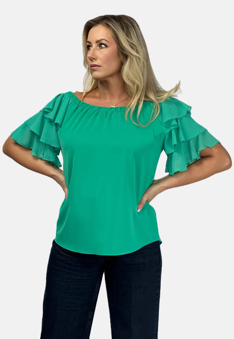 Turkusowa bluzka z dekoltem na ramionach, warstwowo marszczonymi rękawami i gładkim, lekki materiałem. Noszona z ciemnymi dżinsami.