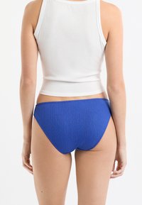 Slip bikini blu a coste con tessuto testurizzato e taglio a vita media, abbinato a una canotta bianca senza maniche.