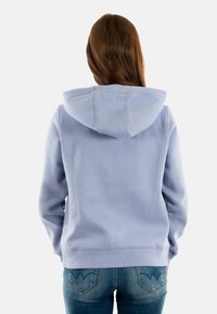 Felpa con cappuccio azzurro chiaro con coulisse, polsini e orlo a coste e una texture morbida. Indossata con jeans aderenti, che mostrano dettagli sulle tasche.