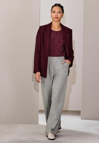 Mujer con cabello trenzado que lleva un blazer y top burdeos, pantalones anchos grises y zapatos blancos, de pie en una habitación de tonos neutros.