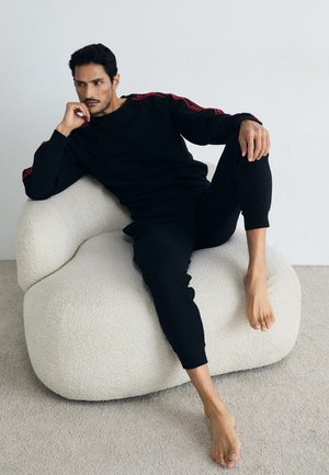 Schwarzes gestricktes Lounge-Set mit roten Akzenten, das eine entspannte Passform und ein geripptes Material aufweist. Modell sitzt auf einem cremefarbenen, plüschigen Sitzsack.