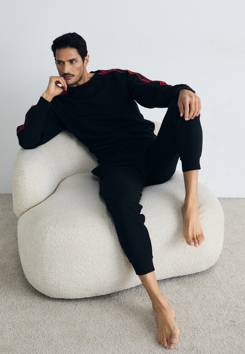 Schwarzes gestricktes Lounge-Set mit roten Akzenten, das eine entspannte Passform und ein geripptes Material aufweist. Modell sitzt auf einem cremefarbenen, plüschigen Sitzsack.