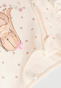 Beige overall med ett brunt kaninmönster och en rosa rosett, omgiven av små blommönster. Materialet verkar mjukt och texturerat.