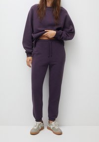 Fioletowy sweatshirt z elastycznymi mankietami, w zestawie z dopasowanymi spodniami dresowymi z elastycznym pasem. Obuwie: szaro-tanowe sneakersy.