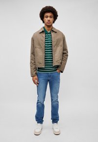 Casaco marrom curto, polo riscado verde e branco, jeans azuis e ténis brancos. Inclui bolsos e tecido texturizado.