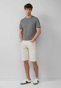 Graues T-Shirt mit kurzen Ärmeln und Brusttasche, kombiniert mit hellcremefarbenen Shorts. Weiße Sneakers mit schwarzen Akzenten runden das Outfit ab.