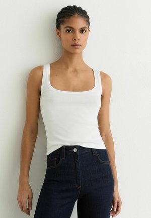 Femeie cu părul împletit, purtând un top alb fără mâneci, cu dungi, și blugi albastru închis, în fața unui perete alb.