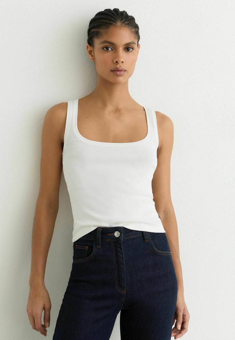 Nainen, jolla on letitetyt hiukset, valkoinen hihaton ribbineulepaita ja tummansiniset farkut valkoista seinää vasten.