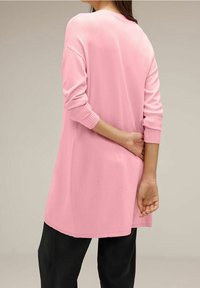 Lång rosa tröja med ribbade muddärmar, rund halsringning och en avslappnad passform, tillsammans med svarta byxor. Mjuk tygkvalitet.