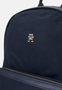 Zaino in nylon blu navy con accessori in tonalità oro, dotato di un logo e di una tasca con zip. Texture liscia con una forma strutturata.