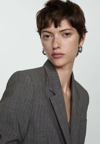 Gray pinstriped blazer with wide lapels, fitted structure, and a pocket detail. Model has short hair and large hoop earrings.