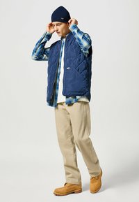 Gilet trapuntato navy sopra una camicia a quadri blu e bianca, pantaloni beige e stivali color beige. Il modello aggiusta un berretto navy. Sfondo neutro.