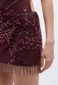 Vīnogu krāsas kleita ar ziedu izšuvumiem, fliteriem un bārkstīm apakšmalā. Caurspīdīgs, teksturēts audums ar izsmalcinātām rotājumiem.