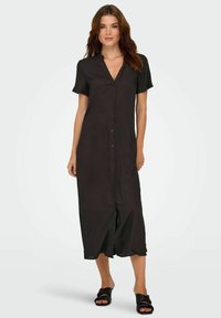 Robe noire à boutons avec des manches courtes, longueur genoux, tissu doux et silhouette légèrement évasée, associée à des sandales noires.