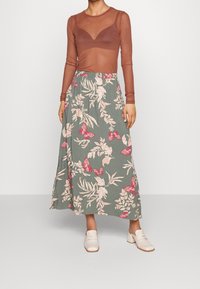 Un haut long à manches transparentes marron avec un décolleté en V, associé à une jupe maxi florale verte ornée de papillons roses et de motifs botaniques.