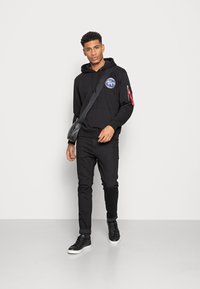 Hoodie preto com um logótipo no peito, bolso com fecho lateral, combinado com jeans pretos e ténis altos pretos. Um visual simples e casual.