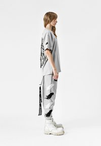 Camisa de gran tamaño en gris con un diseño intrincado de alas en la espalda, combinada con pantalones grises decorados con parches de plumas en blanco y negro y etiquetas, rematada con botas blancas voluminosas.