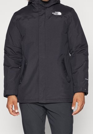 Mężczyzna ubrany w czarną ocieplaną kurtkę z kapturem i logo The North Face, stojący z ramionami swobodnie opuszczonymi wzdłuż ciała.