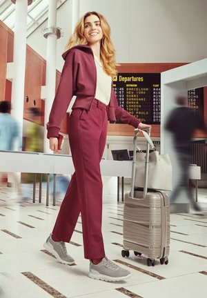 Glimlachende vrouw in bordeauxrode outfit loopt door terminal van luchthaven en trekt een beige koffer met een tas erop, vertrekbord op de achtergrond.
