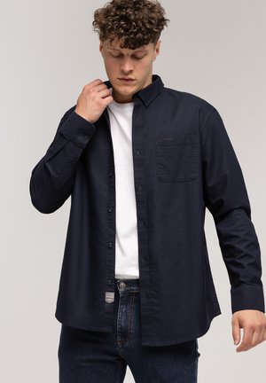 Granatowa koszula z długim rękawem, zapinana na guziki, z kieszenią na piersi i dopasowanym krojem, wykonana z lekkiego materiału, zestawiona z białym T-shirtem.