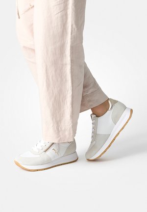 Person wearing light beige pants and white sneakers with beige details and gum soles, standing on a white background.