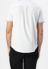 Camicia bianca a maniche corte con dettagli delle maniche in azzurro chiaro e orlo curvo. Tessuto morbido con design minimale e senza chiusure visibili sul retro.