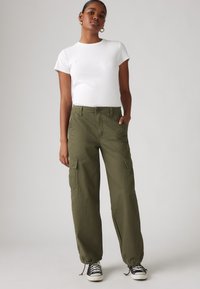 Olivgrüne Cargo-Hosen mit elastischen Bündchen, zwei Seitentaschen und normaler Passform, kombiniert mit einem weißen T-Shirt mit kurzen Ärmeln und schwarzen Turnschuhen.