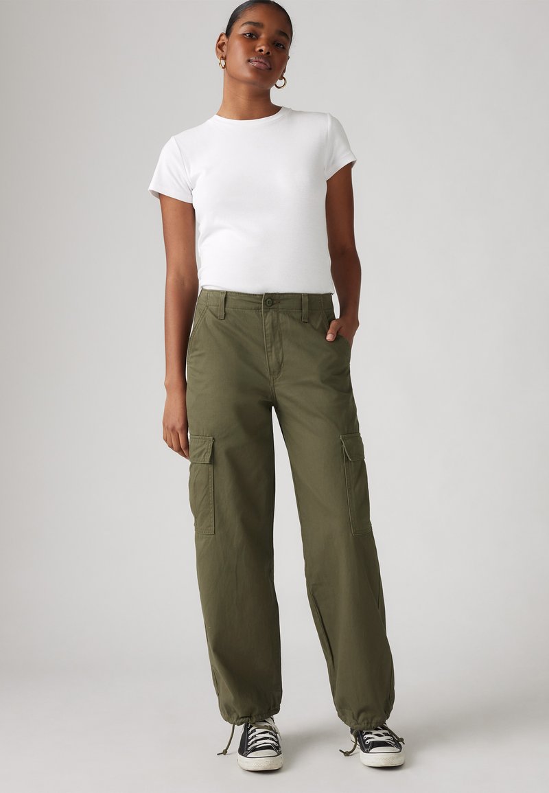 Olivgrüne Cargo-Hosen mit elastischen Bündchen, zwei Seitentaschen und normaler Passform, kombiniert mit einem weißen T-Shirt mit kurzen Ärmeln und schwarzen Turnschuhen.