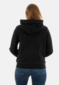 Felpa nera con cappuccio dal morbido tessuto, dotata di tasca a marsupio e polsini a coste. Indossata con jeans aderenti blu. Vista posteriore mostrata.