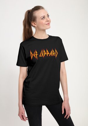 Ung kvinde med brunt hår bundet tilbage, iført sort Def Leppard band t-shirt og sorte bukser, kigger let opad og smiler.