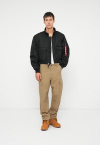 Giubbotto bomber nero con colletto a costine, due tasche frontali e una linguetta rossa sulla cerniera. Indossato con pantaloni cargo beige e stivali color beige.