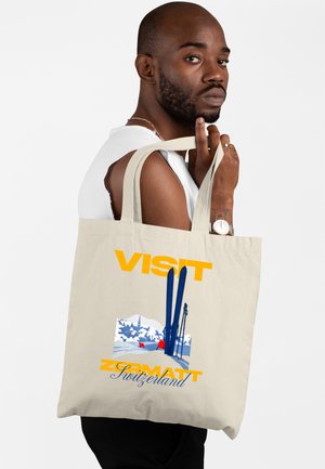 Uomo con barba e orologio tiene una borsa a tracolla con la scritta "Visita Zermatt Svizzera" con grafica di sci e montagne.