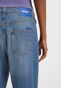 Vue arrière d'une personne portant un jean bleu Hugo avec des patches de logo près de la taille et de la poche, et un haut violet visible au niveau de la taille.
