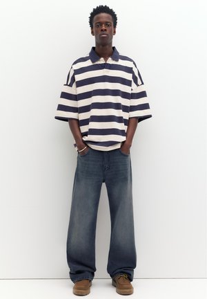 Junger Mann, der an einer weißen Wand steht, trägt ein locker sitzendes marineblau-weiß gestreiftes Hemd, weite blaue Jeans und braune Schuhe.