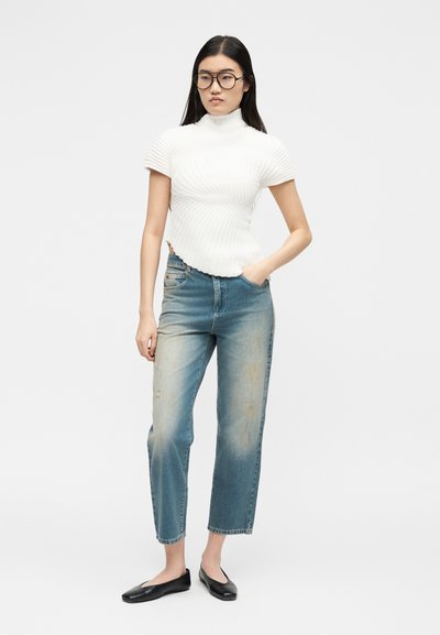 Femme aux longs cheveux noirs et lunettes, portant un haut blanc à manches courtes côtelé et col roulé, un jean bleu délavé et des ballerines noires, debout avec une main dans la poche.