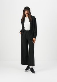 Zwarte cropped jas en wijde broek outfit met een witte blouse. Bevat een opvallend Calvin Klein-logo op de tailleband en elegante zwarte sneakers.