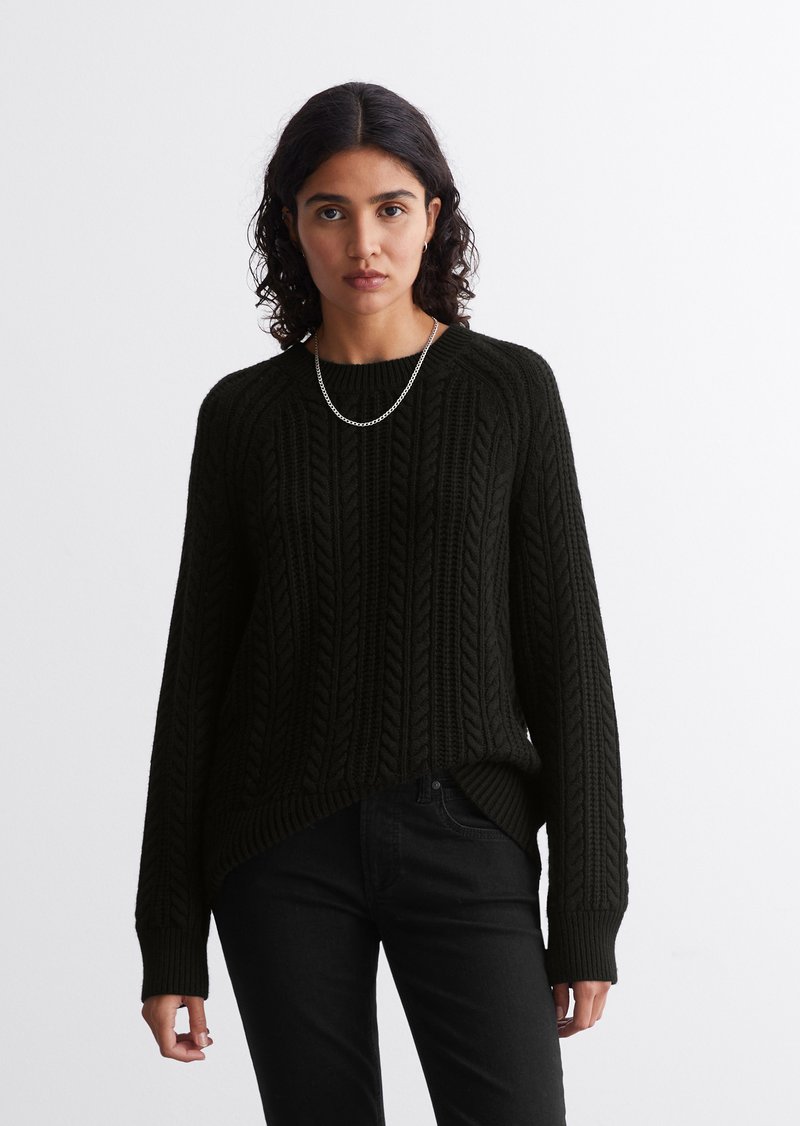 Schwarzer Strickpullover mit strukturiertem Zopfmuster, Rundhalsausschnitt und lockerer Passform. Kombiniert mit schwarzen Jeans für einen monochromen Look.