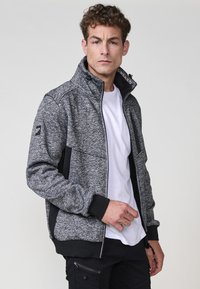 Chaqueta de hombre en gris y negro con cremallera, cuello alto, tejido texturizado, bolsillos laterales y acentos negros en contraste en las mangas y el dobladillo.