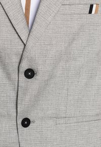 Světlé šedé sako z texturované látky, s černým knoflíkem, jemným károvaným vzorem a proužkovaným detailem na kapse.