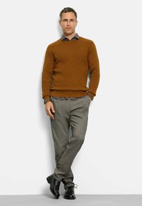 Brauner, gerippter Pullover über einem blumenbedruckten Hemd mit Kragen, kombiniert mit grauen strukturierten Hosen und schwarzen Schuhen; das Model steht mit einer Hand auf der Hüfte.