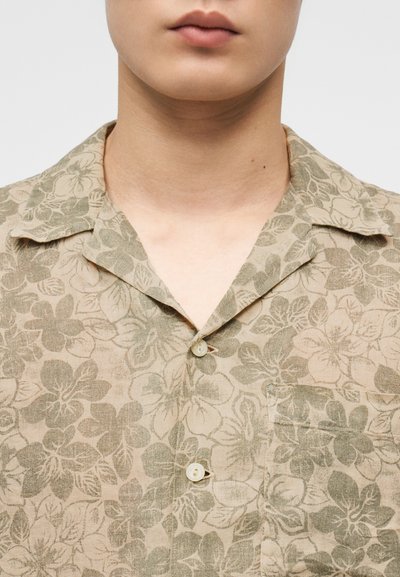 Κοντινό πλάνο του λαιμού και του άνω μέρους του στήθους με πουκάμισο μπεζ με πράσινο λουλουδένιο σχέδιο και μπροστινή τσέπη, με κλεισμένα δύο κουμπιά.