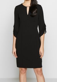 Svart klänning med trekvartslånga veckade ärmar, nyckelhålsutringning, slät tyg. Rak skärning med minimal detaljering, lämplig för formella tillfällen.