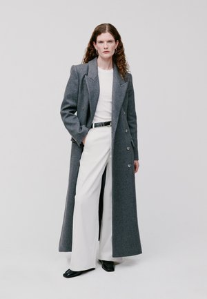 Long manteau en laine gris avec un design à double boutonnage, porté sur un haut blanc et un pantalon blanc large, avec des accessoires noirs et des chaussures plates.