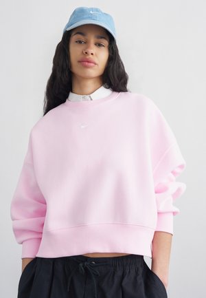 Frau trägt ein hellrosa Nike-Sweatshirt, eine blau-jeansfarbene Nike-Kappe, schwarze Hose, mit den Händen in den Taschen, stehend vor einem weißen Hintergrund.