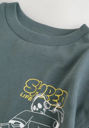 Tamsiai žalia marškinėliai suapvalintu kaklu ir geltonu užrašu „SHRED LIFE“ bei baltu grafiniu vaizdu - kaukolė su ausinėmis.