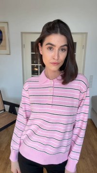 Kvinne med rett mørkt hår iført en rosa stripete polotrøye med lange ermer, stående innendørs i et rom med lyse vegger.