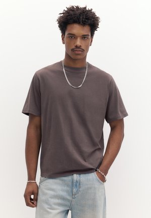 Tânăr cu păr creț, purtând un tricou maro, lanț argintiu la gât, brățări argintii și blugi deschiși la culoare, stând în picioare pe un fundal alb.