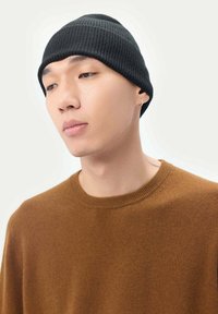 Svart ribbad mössa, snug passform, mjukt material. Bärs tillsammans med en brun tröja med rundhals. Enkel design utan synliga logotyper.