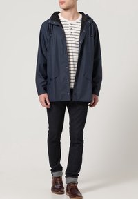 Giovane uomo con una giacca a vento blu scuro sopra una maglietta a righe, abbinata a jeans scuri e stivali marroni, in posa su uno sfondo neutro.